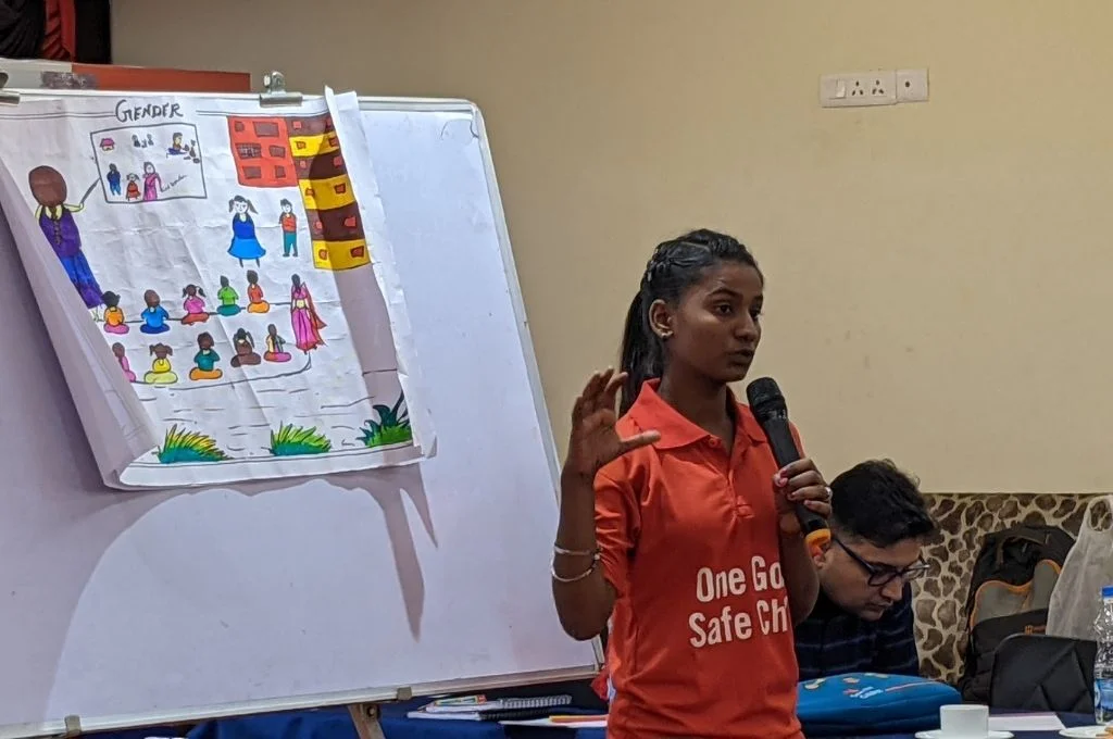 लाल रंग की टीशर्ट में हाथ में माइक लिए हुए एक लड़की दर्शकों से बात कर रही है। उसके पीछे एक सफ़ेद बोर्ड है। लूसी शर्मा लैंगिक समानता पर कार्यशाला का आयोजन करती है और लोगों को प्रशिक्षण देती है_युवा क्रिकेटर