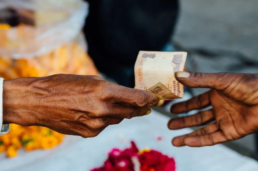 फूलों की धून्धली पृष्ठभूमि के साथ एक हाथ से दूसरे हाथ में दस रुपए के एक नोट का आदान-प्रदान किया जा रहा है-क्राउडफंडिंग टैक्स