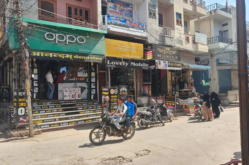 एक सड़क पर चलती मोटरसाइकिल और किनारे मोबाइल और लैपटॉप रिपेयरिंग की दुकानें_ ई-कचरा