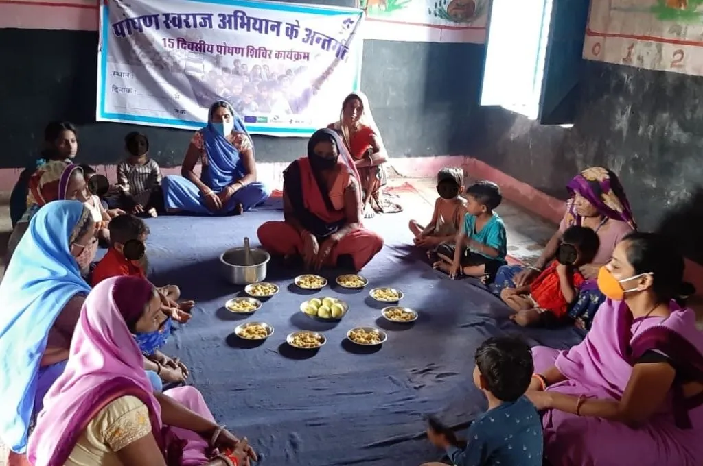 एक कमरे में कुछ महिलायें और बच्चे बैठकर खाना खा रहे हैं_पोषण